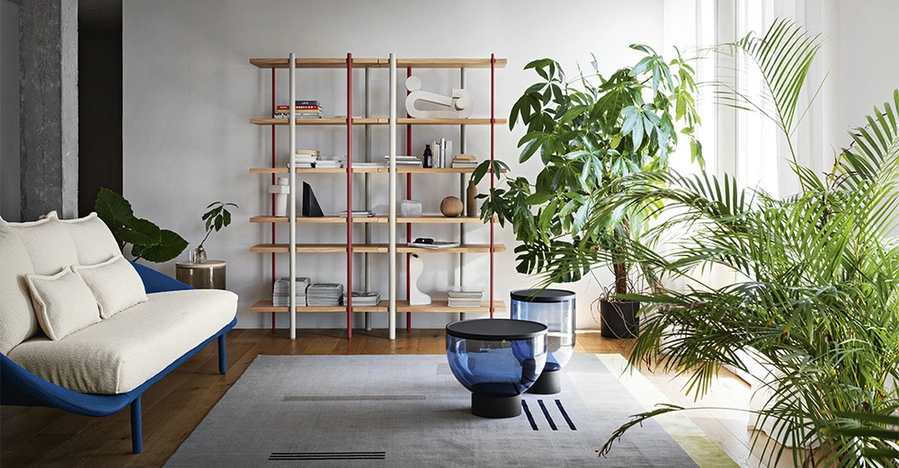 Sofa steht vor Bücherregal in einer modernen, hellen Wohnung mit Holzboden umgeben von großen grünen Zimmerpflanzen und zwei blauen Glastischen auf einem Teppich.