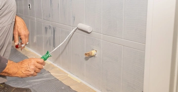 älterer Mann überstreicht die alten Fliesen im Bad mit grauer Fliesenfarbe. Kleiner Rollpinsel als Werkzeug. Armaturen und Auslässe alle fachgerecht mit Malerkrepp abgeklebt. 