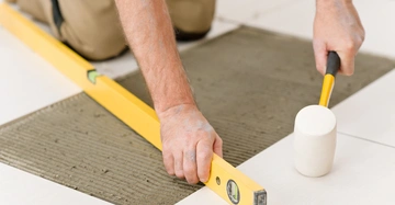 Hande richten mit Wasserwaage und Gummihammer Fliesen auf feuchtem Zement aus Knieende Person im Hintergrund helle Umgebung