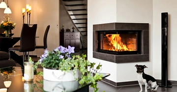 Hund sitzt ruhig neben brennendem Kamin in modernem Wohnzimmer nahe einem Tisch mit Blumenarrangement und einem Essbereich mit Stühlen. Eine Treppe ist im Hintergrund sichtbar.