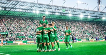 Fußballspieler umarmen sich auf einem Fußballfeld, umgeben von jubelnden Zuschauern in einem vollbesetzten Stadion. Die Spieler tragen grüne Trikots mit Logos wie Allianz RHIEM ENERGIE auf der Vorderseite.