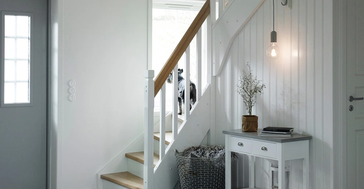 Tür steht still in einem ruhigen, hellen Flur mit einer Treppe, ein Hund steht und blickt durch das Fenster in den Hof, ein Tischchen mit Dekoration steht daneben.