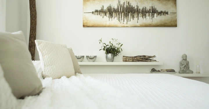 Galerie-Bild 5: Ein großes Bett mit hellen Kissen steht in einem minimalistisch eingerichteten Schlafzimmer. An der weißen Wand hängt ein abstraktes Gemälde. Ein Regal mit Dekorationen befindet sich dahinter.