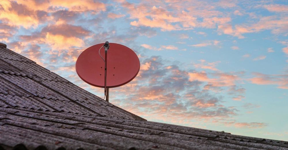 Satellitenschüssel stehend auf einem geneigten Dach vor einem Himmel mit orange und blau gefärbten Wolken in der Abenddämmerung