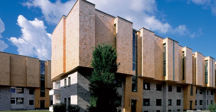 Galerie-Bild 1: Ein modernes Wohngebäude mit hölzerner und steinerner Fassade steht unter einem blauen Himmel mit wenigen Wolken in einem grünen, offenen Bereich.