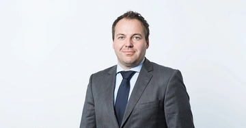 Ein Mann trägt einen grauen Anzug mit blauem Hemd und Krawatte und steht vor einem weißen Hintergrund.