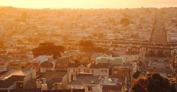Viele Gebäude stehen still in der Abendsonne in einer weitläufigen städtischen Gegend die sich bis zum Horizont erstreckt mit einer langen geraden Straße die durch die Stadt führt.