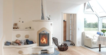 Kind liegt auf einer gepolsterten Bank neben einem Kamin mit brennendem Feuer in einem hellen, gemütlichen Wohnzimmer mit Holzboden und großen Fenstern, die viel natürliches Licht hereinlassen.