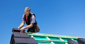 Handwerker installiert schwarze Dachziegel auf einem halb fertigen Dach; er arbeitet konzentriert und trägt Werkzeug am Gürtel; klarer blauer Himmel im Hintergrund.