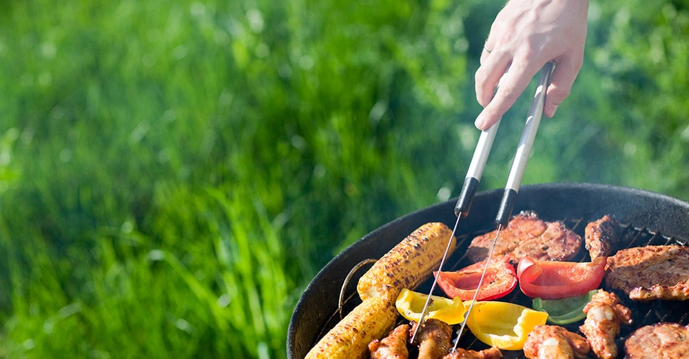 Hand mit Grillzange wendet Grillgut auf dem Grill im grünen Garten.