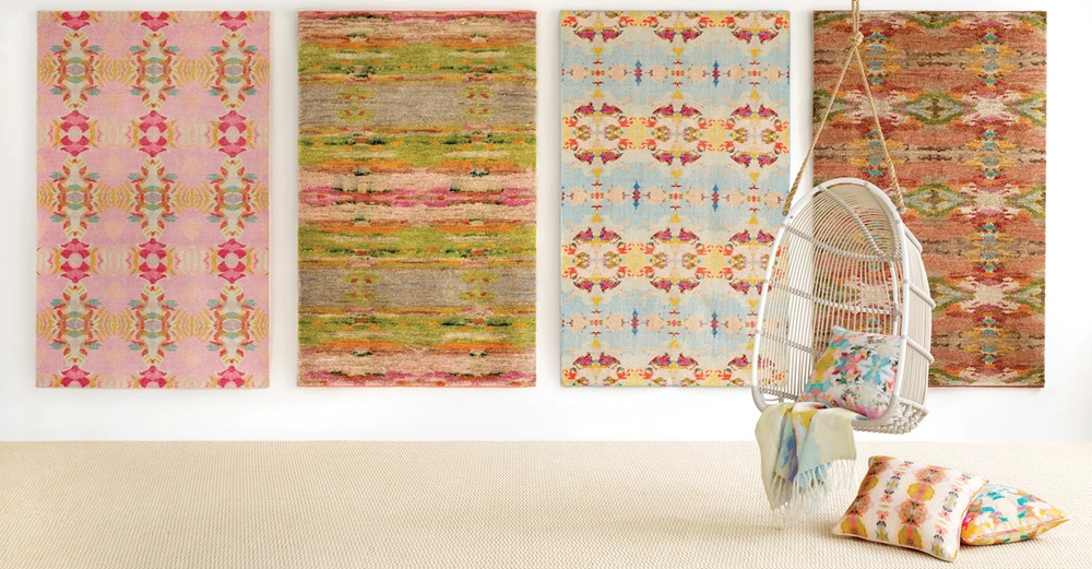 Vier bunte, gemusterte Teppiche hängen an einer Wand in einem hellen Raum vor einer beigen Bodenmatte links während ein weißer Hängesessel mit bunten Kissen rechts hängt.