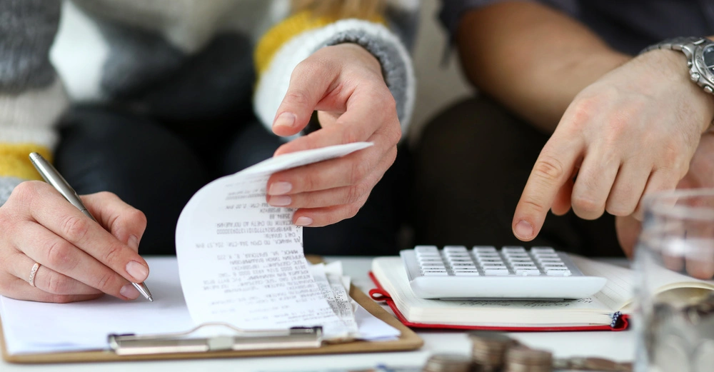 Eine Person hält einen Stift und einen Kassenzettel in der Hand während eine andere Person auf einen Taschenrechner zeigt im Hintergrund sind ein Clipboard Notizbuch und Münzen zu sehen