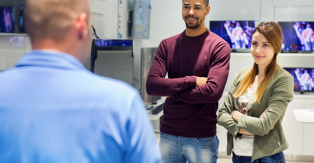 Zwei Personen stehen mit verschränkten Armen vor einem Verkäufer in einem Geschäft mit Fernsehern an den Wänden im Hintergrund.
