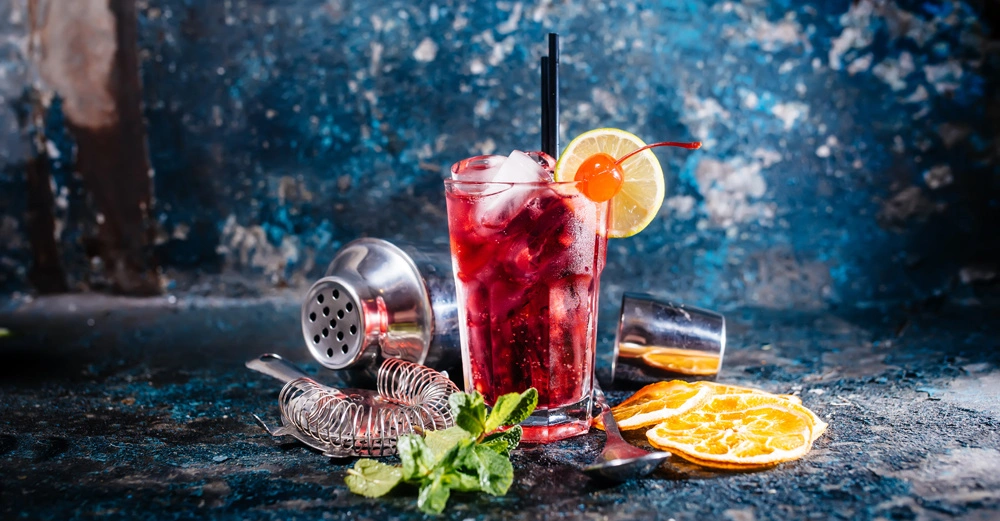 Ein Glas mit rotem Getränk und Eiswürfeln steht neben einer Zitronenscheibe und Kirsche auf einem metallischen Untergrund umgeben von Barzubehör und Orangenscheiben vor einem rustikal blauen Hintergrund.