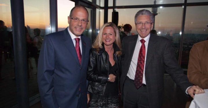 Galerie-Bild 59: Drei Personen stehen lächelnd in einem modernen Raum mit Glaswänden und Blick auf den Sonnenuntergang zwei Männer in Anzügen und eine Frau in einer Lederjacke und einem roten Rock.