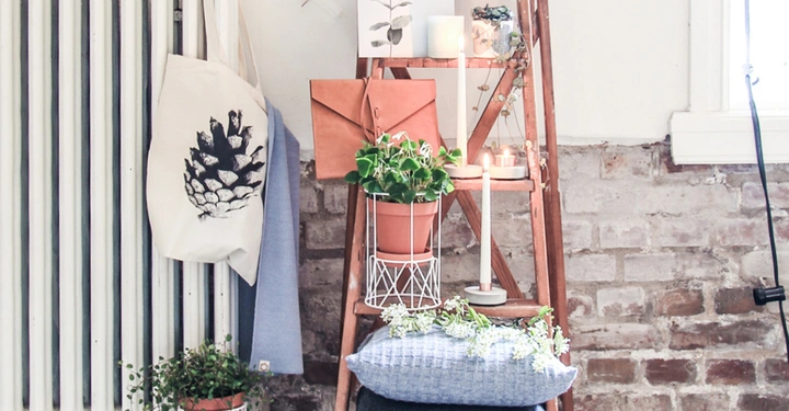 Ein Holzgestell trägt Pflanzen Töpfe und Kissen in einem rustikalen Raum mit Ziegelwand und schwarz-weißen Fliesen Heizkörper an der linken Wand. Stofftasche hängt links.