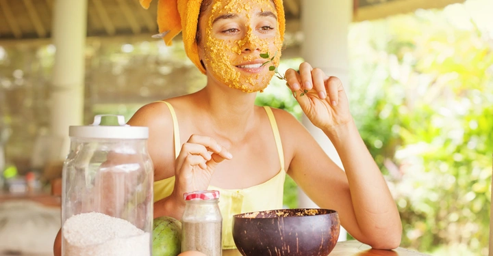 Frau mit orangefarbenem Handtuch auf dem Kopf trägt eine Gesichtsmaske auf und hält Kräuter in der Hand in tropischem Outdoor-Umfeld mit Zutaten wie Reis und Eiern auf einem Tisch