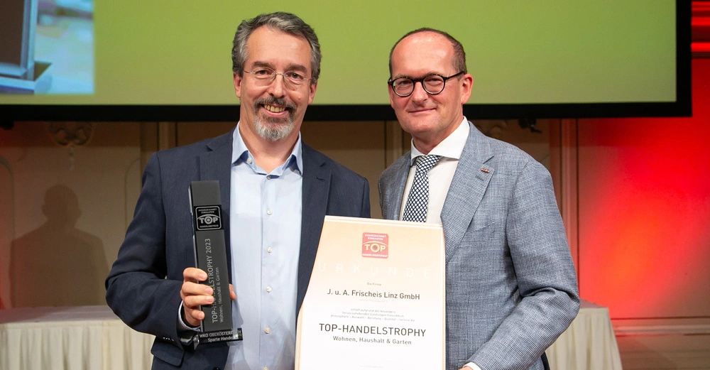 Zwei Männer halten eine Urkunde mit dem Text J. u. A. Frischeis Linz GmbH und einen Preis in einem Innenraum mit roter Beleuchtung und einem großen Bildschirm im Hintergrund.
