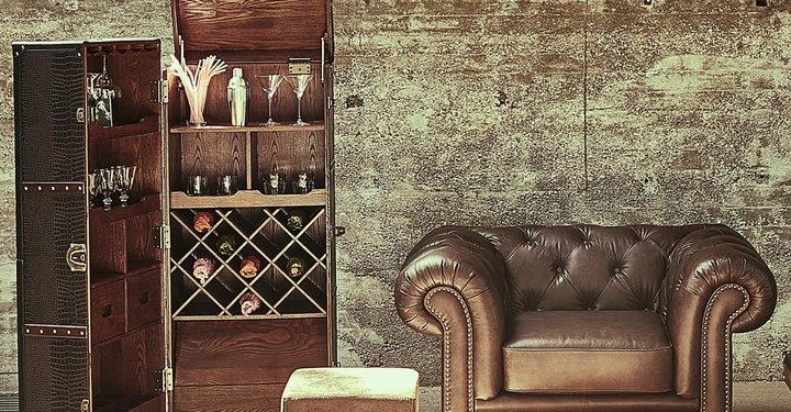 Holzschrank mit Gläsern und Weinflaschen steht offen neben Ledersessel und Hocker in einem rustikalen Raum mit Steinwand.