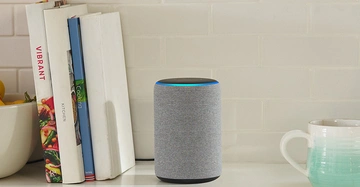 Ein zylindrischer Lautsprecher mit leuchtendem Ring oben steht auf einer Küchentheke neben drei Kochbüchern (VIBRANT KITCHEN und Cooking) und einer grün-weißen Tasse.