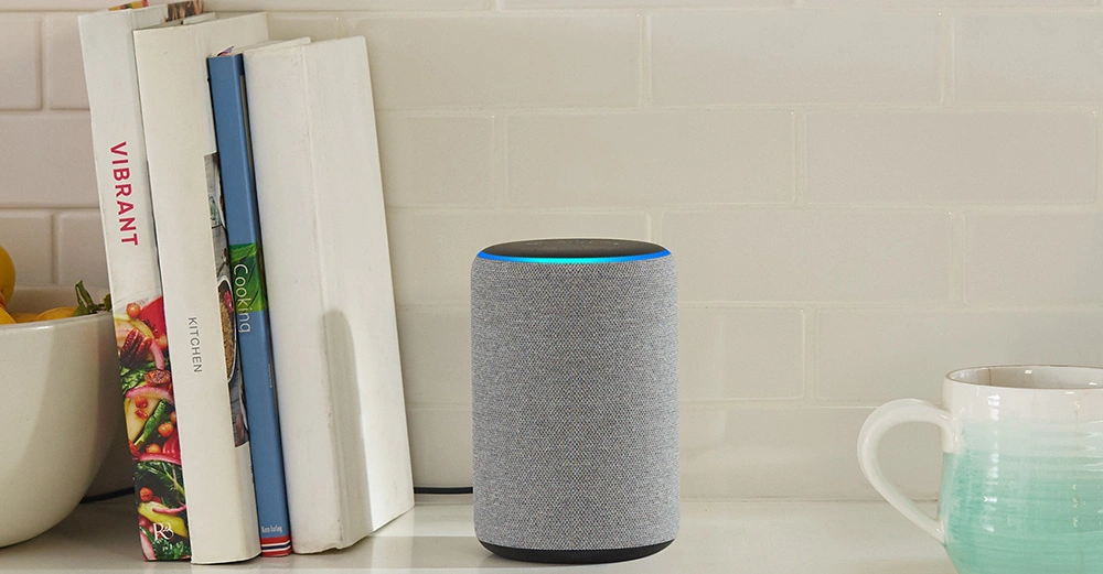 Ein zylindrischer Lautsprecher mit leuchtendem Ring oben steht auf einer Küchentheke neben drei Kochbüchern (VIBRANT KITCHEN und Cooking) und einer grün-weißen Tasse.