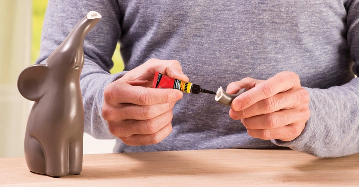 Eine graue Elefantenfigur wird mit Klebstoff an einem abgebrochenen Rüssel repariert Hände im Vordergrund halten den Rüssel und eine Tube UHU Kleber Holztisch im Innenraum