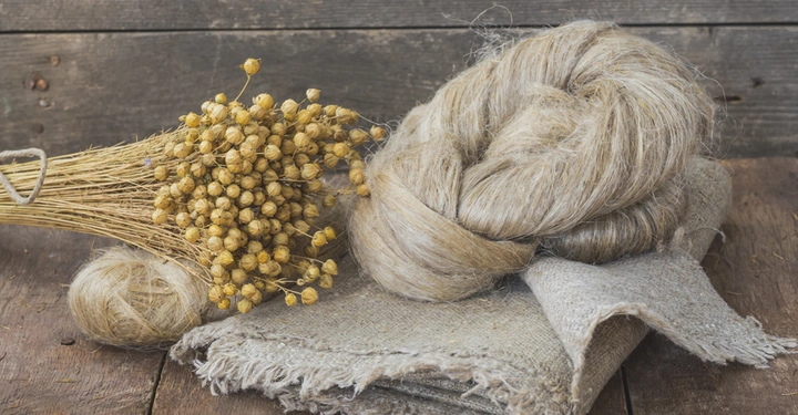 Ein Bündel getrockneter Pflanzen ruht auf einer Holzoberfläche neben einem Haufen Leinenfasern und grobem Stoff in einem rustikalen Raum.