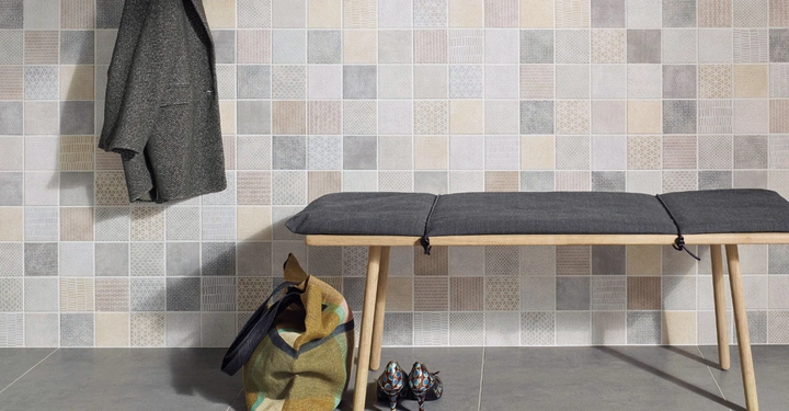 Gepolsterte Holzbank steht vor einer Wand mit gemusterten Fliesen. Eine Jacke hängt neben einem Hut an der Wand. Auf dem Boden steht eine bunte Tasche und zwei Schuhe