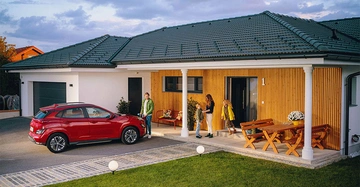 Ein rotes Auto parkt vor einem modernen Einfamilienhaus Familieneinheit steht auf der Veranda der Vater hält einen Jungen an der Hand während zwei Frauen neben einem Holztisch stehen.