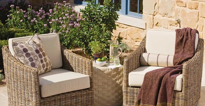 Zwei Rattanstühle mit Kissen stehen auf einer Terrasse vor einem Steinhaus und einem Garten mit blühenden Pflanzen. Ein Beistelltisch dazwischen hat Krüge mit Getränken und Limetten.