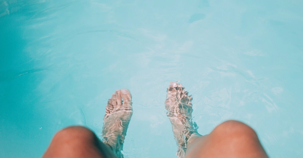 Füße tauchen ins klare blaue Wasser eines Pools ein umgeben von der Ruhe einer Wasserlandschaft.