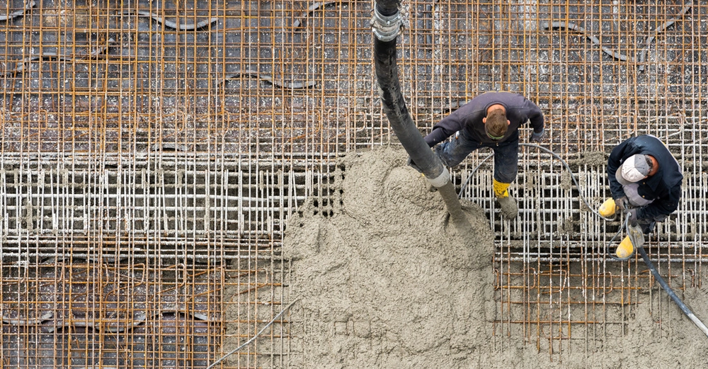 Zwei Bauarbeiter gießen mit einem Schlauch nassen Beton auf die Stahlbewehrung unter ihnen auf.