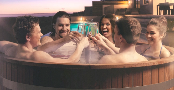 Galerie-Bild 8: Fünf Personen sitzen in einem runden, hölzernen Whirlpool draußen und stoßen mit Sektgläsern an. Im Hintergrund sind moderne Häuser und ein sonniger Himmel bei Sonnenuntergang zu sehen.