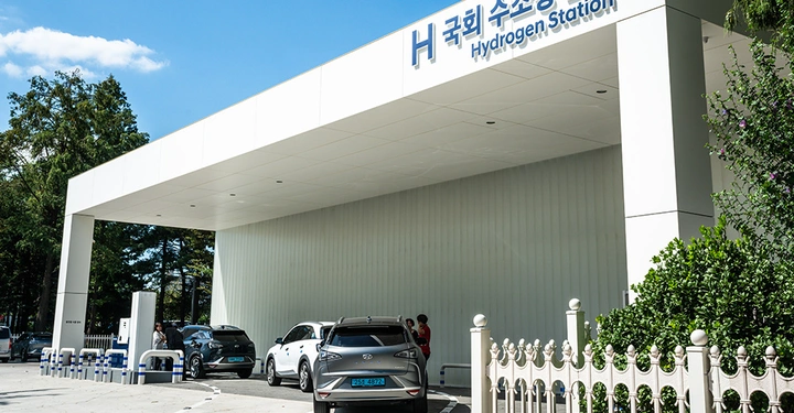 Autos tanken an einer Wasserstofftankstelle unter einem überdachten Bereich im Freien mit Grünflächen im Hintergrund. Beschilderung lautet Hydrogen Station.