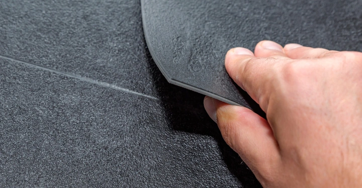 Hand hebt eine Ecke eines dunklen Bodenbelags in Nahaufnahme an um die Textur zu zeigen. Bodenstruktur ist rau und dunkel.