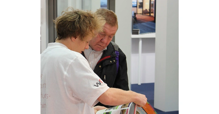 Galerie-Bild 18: Eine Person in einem weißen T-Shirt zeigt einer anderen Person in einer Jacke etwas in einem offenen Buch in einem modernen Raum mit weißen Wänden und blauen Akzenten.