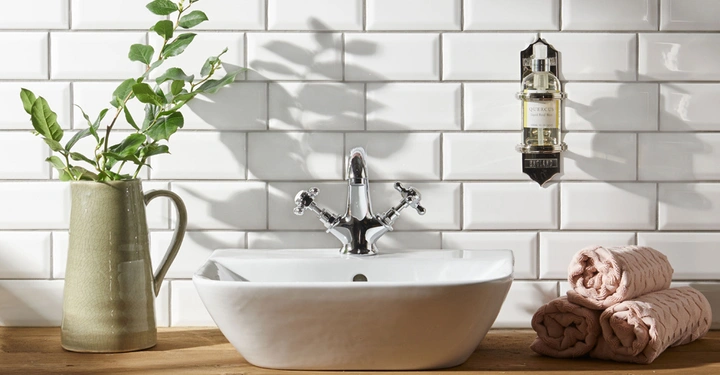 Waschbecken steht auf Holzoberfläche Wasserhahn in der Mitte weiß geflieste Wand Vase mit grünen Zweigen links drei gerollte rosa Handtücher rechts Wandspender mit Text Quercus sensuous cologne Penhaligon’s