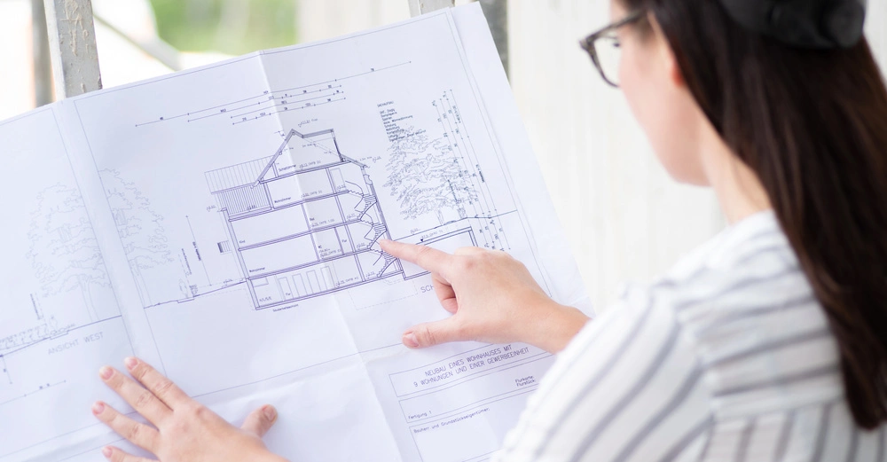 Person zeigt auf Bauplan eines Mehrfamilienhauses mit 9 Wohnungen und einer Gewerbeeinheit im Freien. Draussen im Hintergrund sind verschwommene Bäume sichtbar. Text: Neubau eines Wohnhauses mit 9 Wohnungen und einer Gewerbeeinheit.