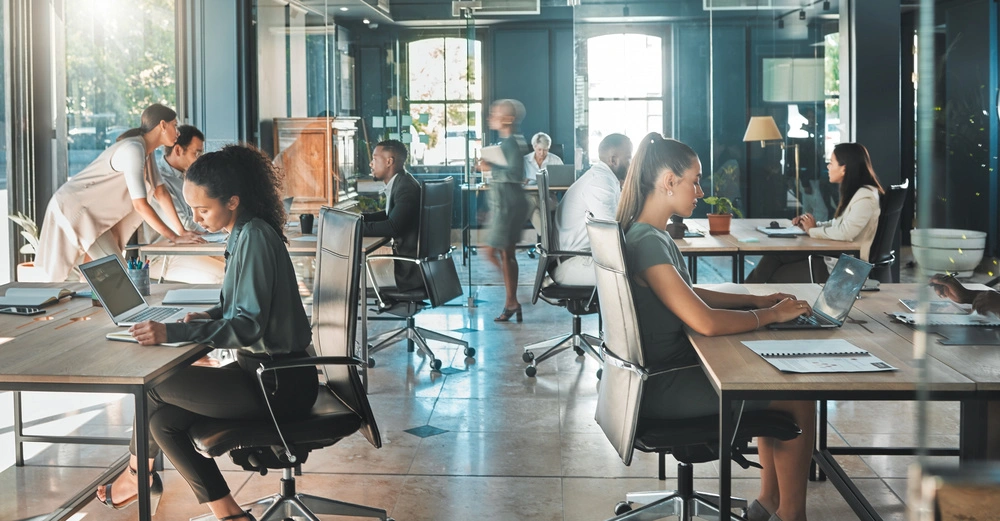 Menschen arbeiten an Computern und Schreibtischen in einem modernen, lichtdurchfluteten Büro mit großen Fenstern und Glaswänden und leuchtendes Grün ist draußen zu sehen. Sie sitzen, tippen und stehen.