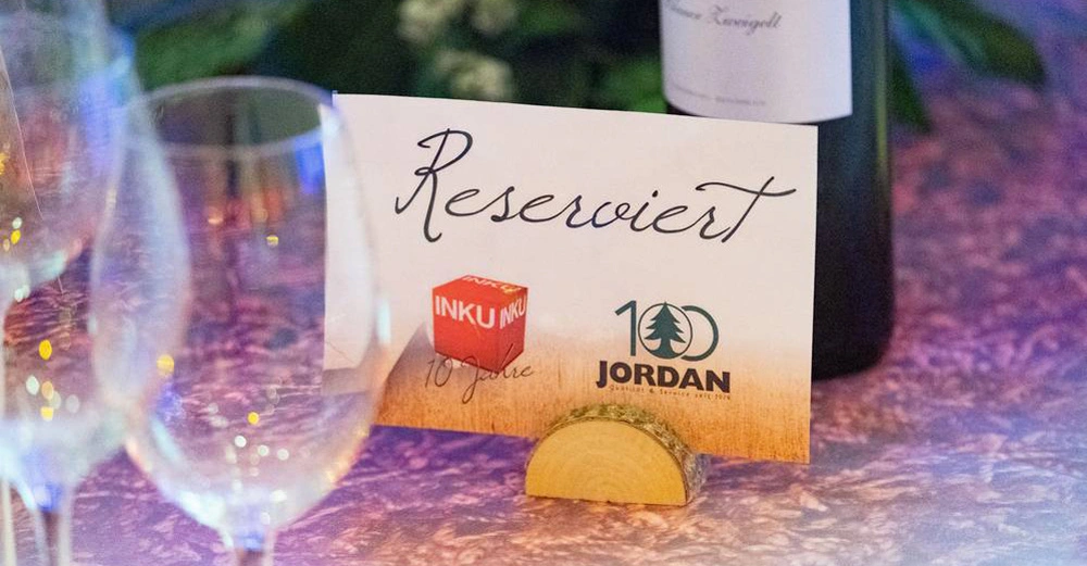 Ein Reserviert-Schild steht auf einem Holztisch Halterung aus Baumstamm umgeben von Weingläsern und einer Weinflasche darauf ist ein Würfel mit der Aufschrift INKU und der Text INKU 10 Jahre und 100 JORDAN.