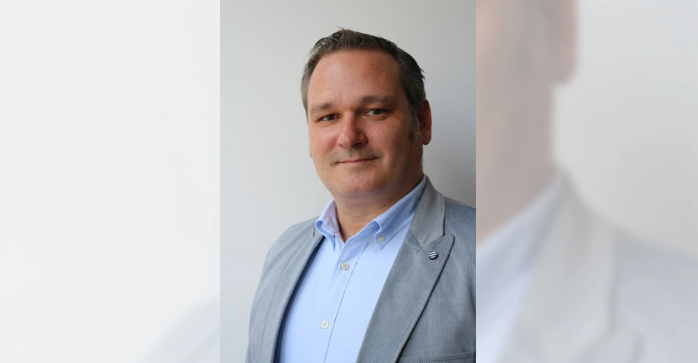 Mann trägt einen hellgrauen Anzug und ein hellblaues Hemd lächelt leicht stehend vor einer weißen Wand in Innenraum.