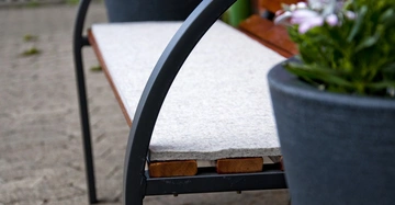 Bank mit grauem Polster bespannt steht auf einem gepflasterten Boden mit Pflanze im Vordergrund daneben befindet sich ein großer Blumentopf mit Blumen rechts im Hintergrund weitere Gartenmöbel