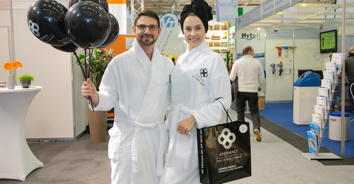 Galerie-Bild 5: Ein Mann und eine Frau in weißen Bademänteln stehen in einer Messehalle. Der Mann hält schwarze Luftballons. Die Frau trägt eine Tasche mit der Aufschrift „elements“. Text im Bild: "BadRenovierung" "elements".