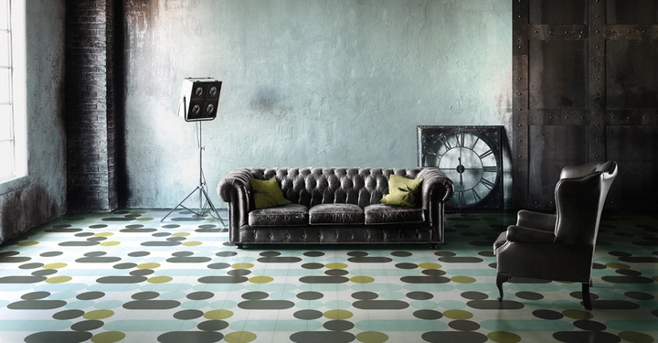 Galerie-Bild 6: Ein graues Ledersofa mit grünen Kissen steht vor einer rustikalen Wand Viktorianischer Sessel rechts Retro-Stehlampe links ein großes Zifferblatt im Hintergrund farbenfroher gemusterter Boden in einem industriellen Raum.