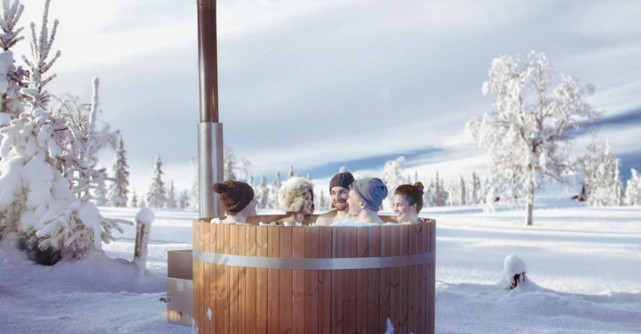 Galerie-Bild 1: Mehrere Menschen sitzen in einem Holzbadezuber im Schnee sie tragen Mützen umgeben von einer verschneiten Winterlandschaft mit Bäumen und klarem Himmel im Hintergrund