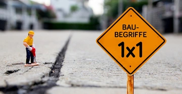 Ein Spielzeugbauarbeiter verwendet einen Presslufthammer in einem Betonriss auf einem Bürgersteig. Ein gelbes Baustellenschild daneben trägt den Text BAU-BEGRIFFE 1x1 mit verschwommenen Gebäuden im Hintergrund