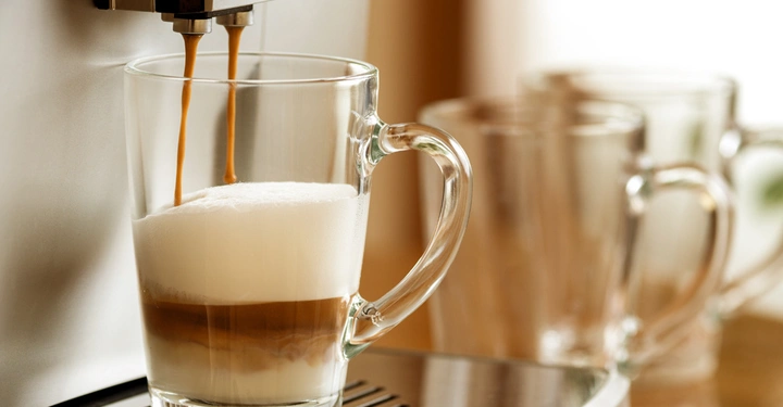 Ein halbvolles Glas wird von einer Kaffeemaschine mit Schaum und Kaffee gefüllt im Hintergrund stehen zwei weitere leere Gläser auf einem metallenen Tablett.