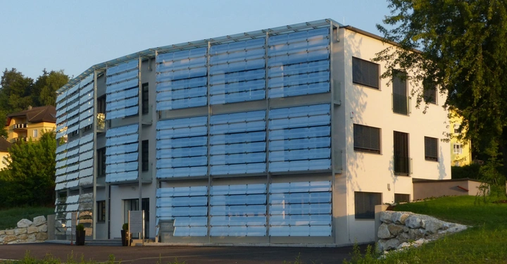 Ein modernes Gebäude mit großen horizontalen Lamellen vor den Fenstern. Es steht in einer grünen Umgebung neben anderen Häusern und ist von Bäumen umgeben.