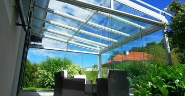 Gartenmöbel aus Rattan stehen unter einem Glasdach in einem Garten vor einem Haus mit roten Dächern und grüner Vegetation im Hintergrund ein klarer blauer Himmel darüber