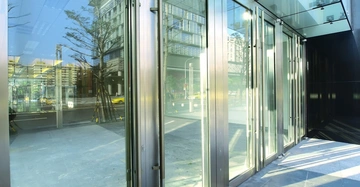 Glas-Eingangstüren reflektieren ein modernes Stadtbild mit Gebäuden und einem vorbeifahrenden gelben Taxi draußen bei Tageslicht. Der Boden besteht aus grauen Fliesen.